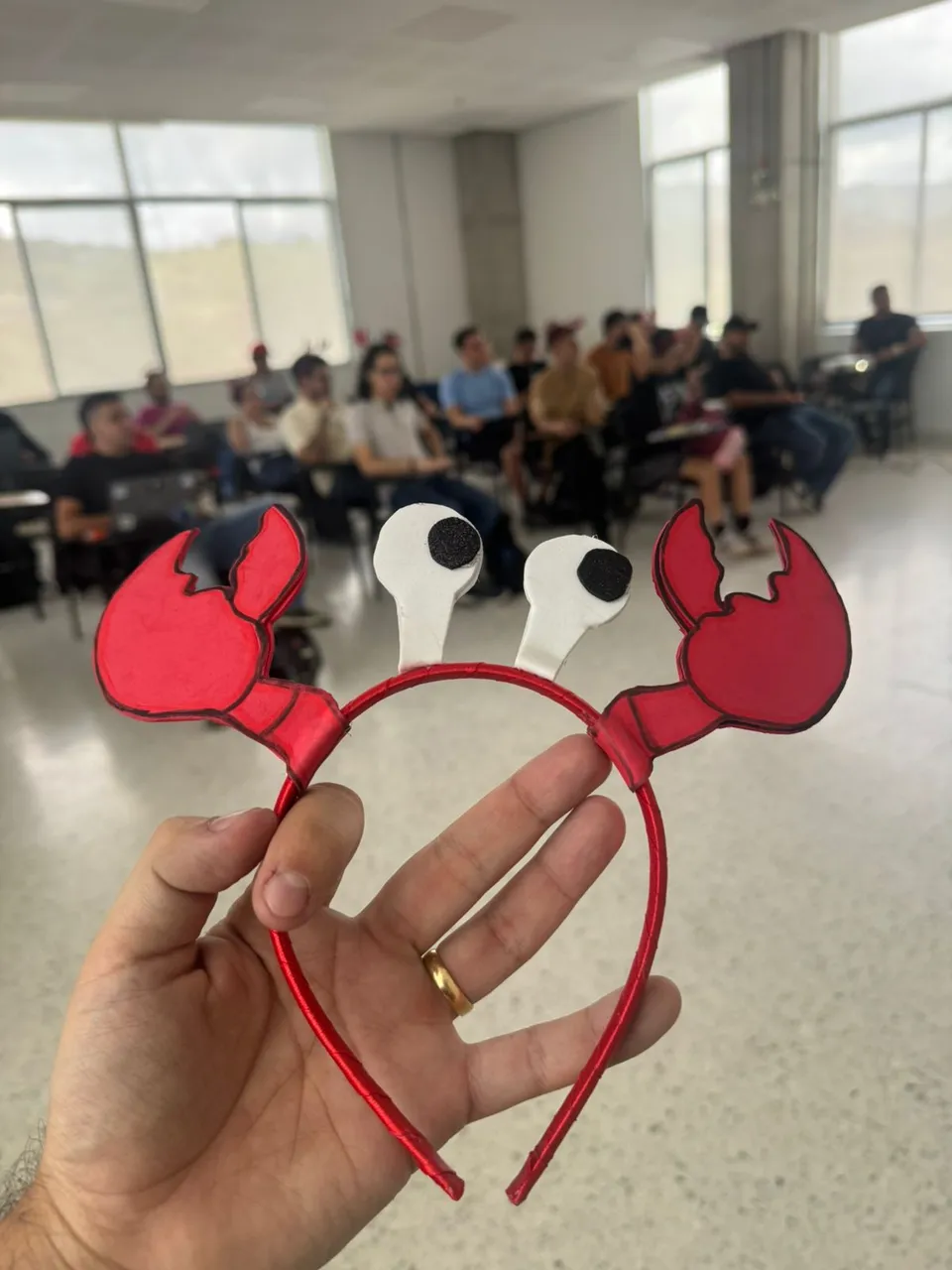 Mano sosteniendo una diadema con tenazas rojas de langosta, la mascota Moltys, con el público sentado detrás